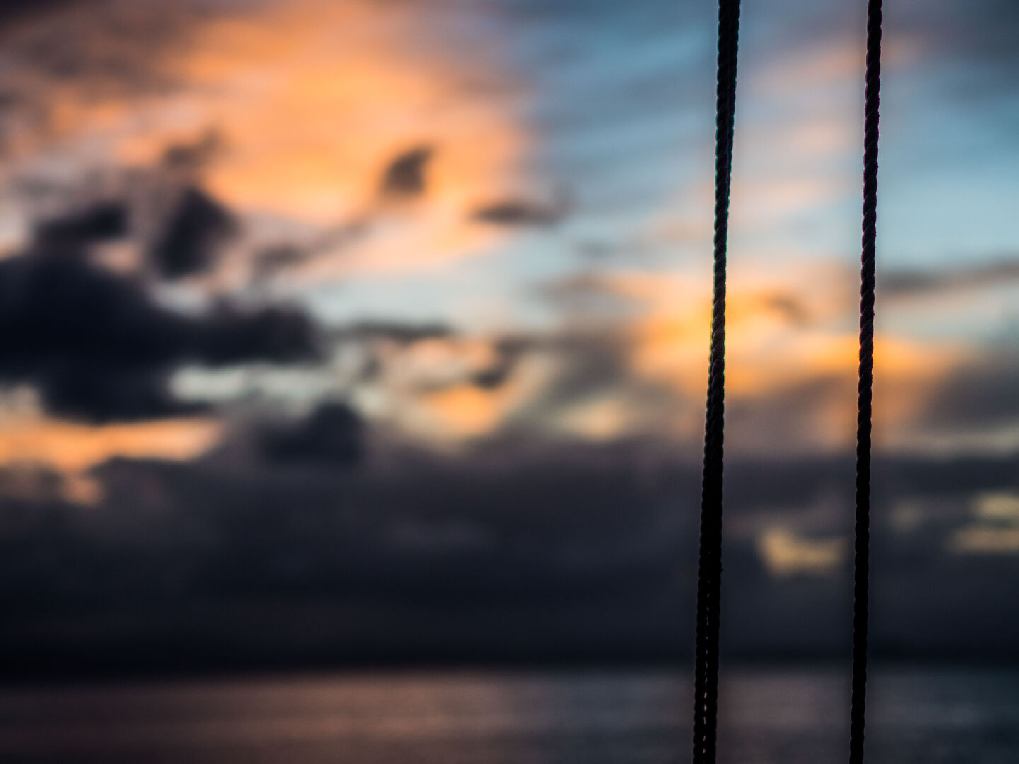 The sun is setting behind clouds in the distance, colouring them partly orange, with two ropes from the sailing boat running through the image from top to bottom