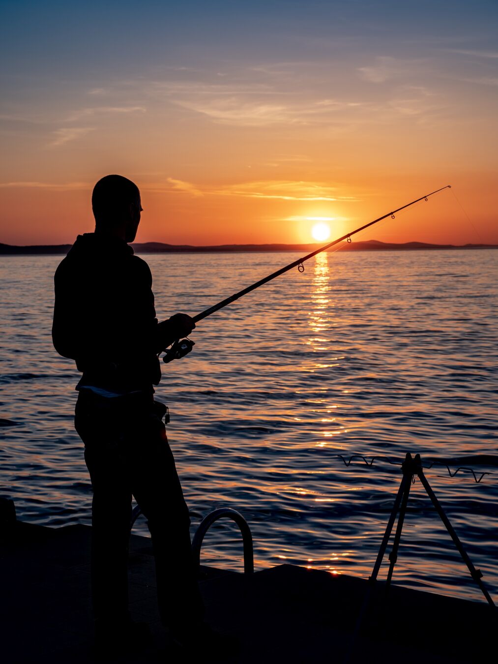 A man fishing while behind him the sun is setting into the sea, setting the sky on fire