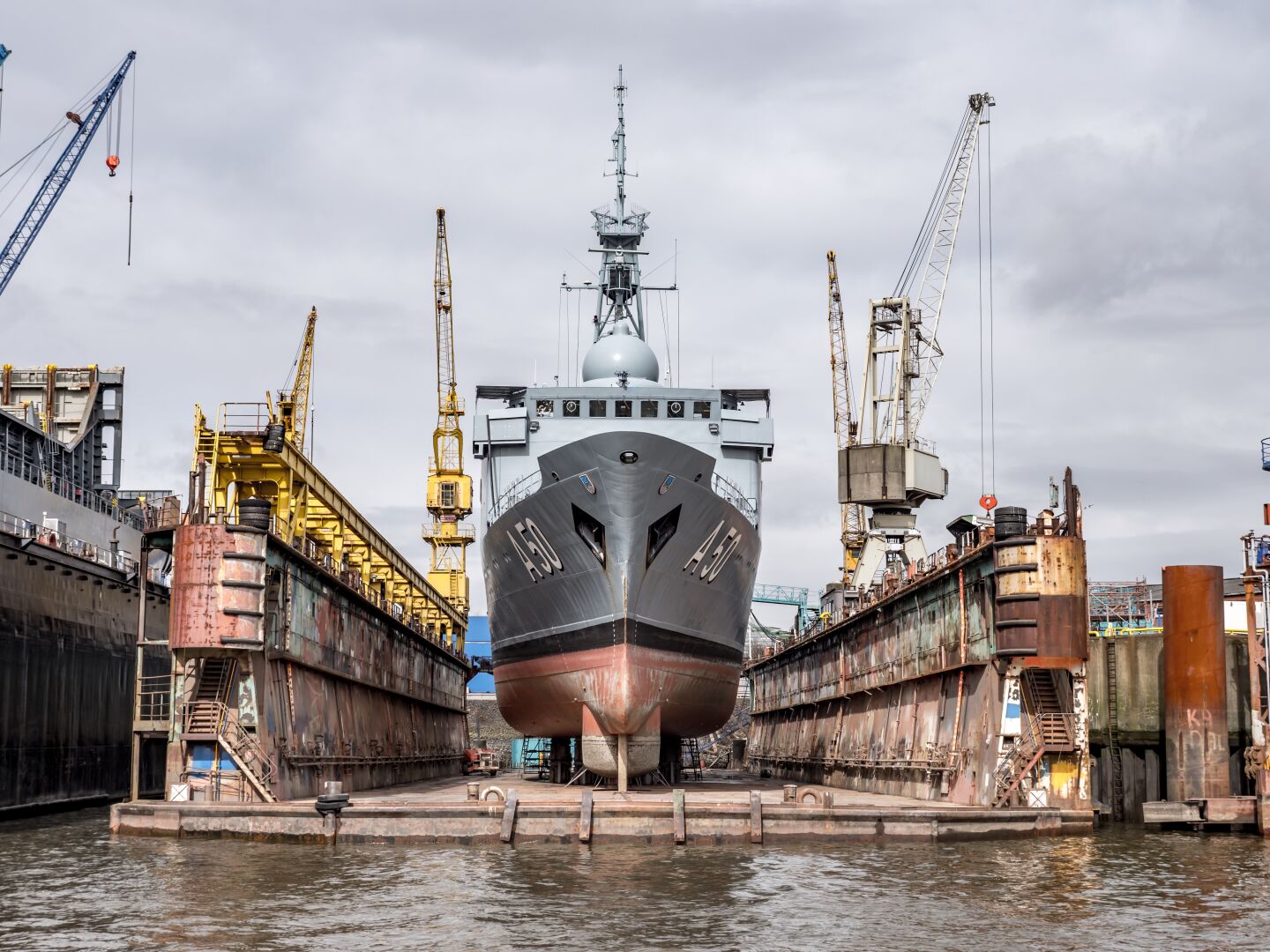 A shipyard with a blue military ship in it, cranes left and right, has pushed the ship out of the water, so it’s while form is visible