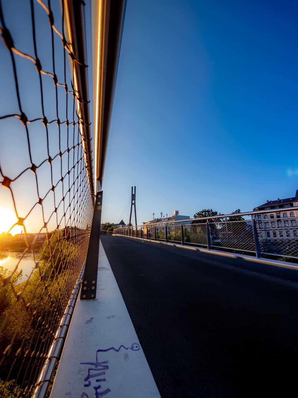 The sun is setting on the left, shining through the wire frame of the railing of the bridge I am standing on. In the distance the city is continuing