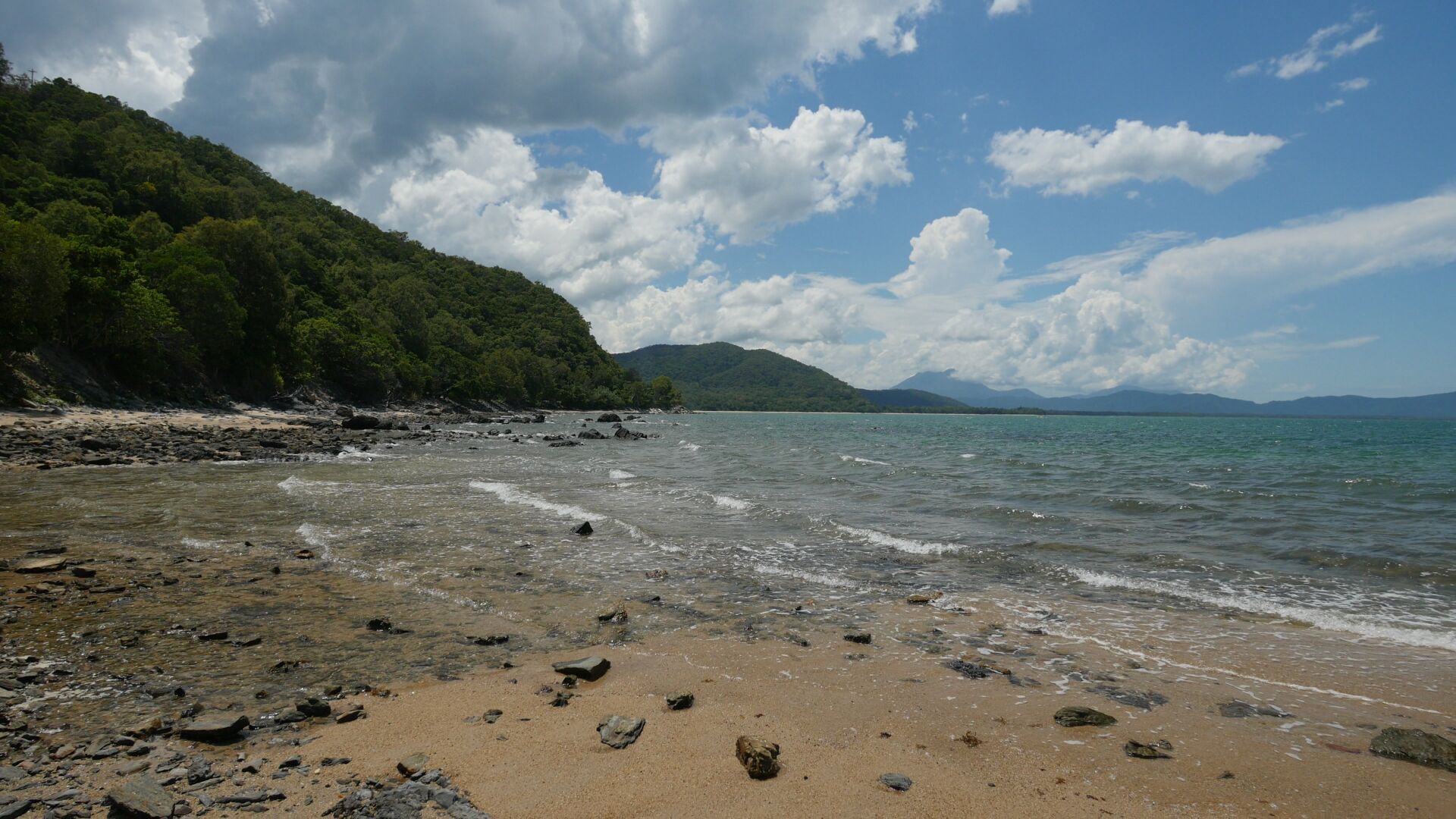 A beach covered with smaller and bigger rocks, with the blue sea on the right and a forest coast on the left side