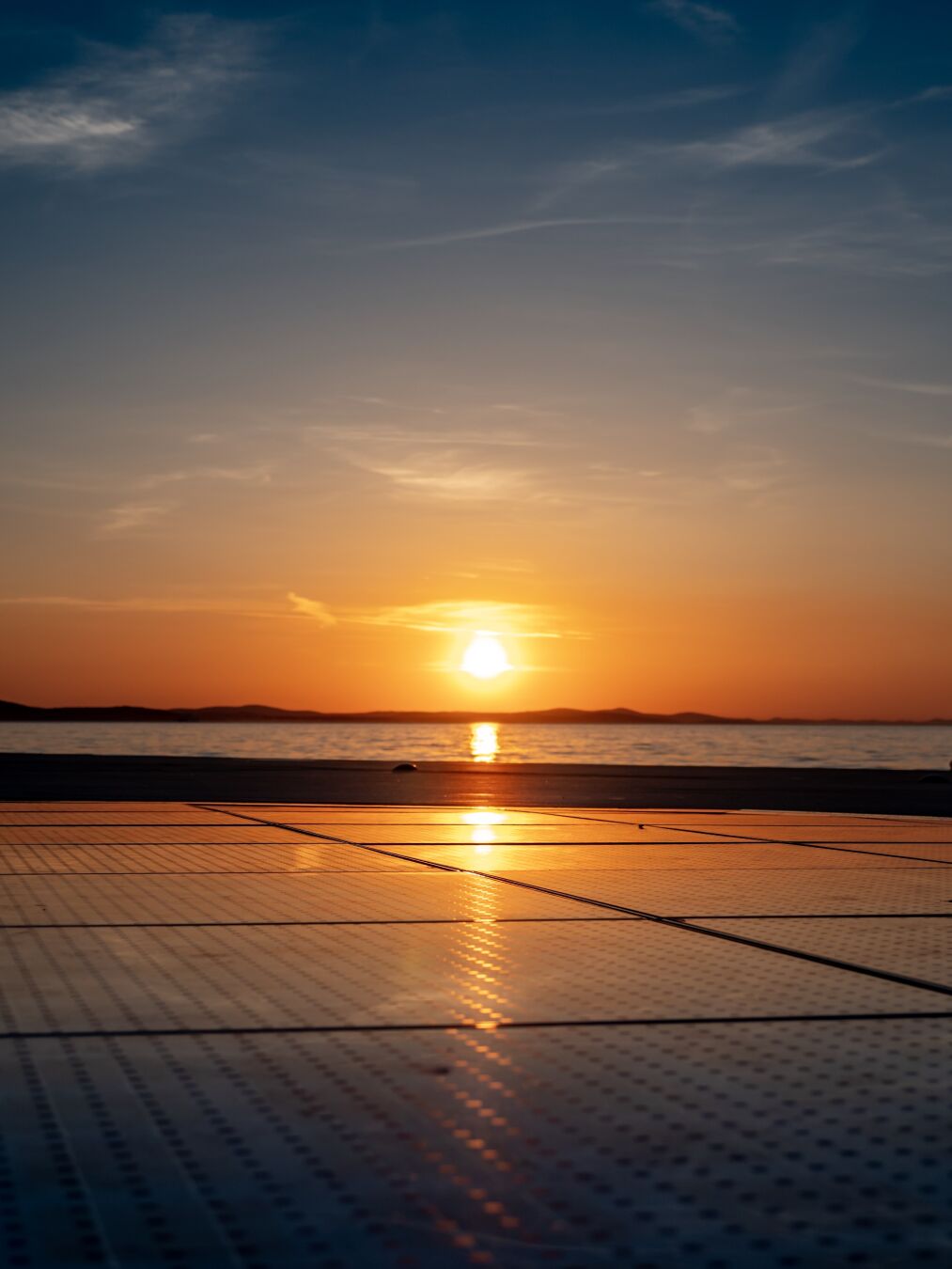 The sun is setting in the background, with its golden light mirrored in the sky, on the sea and the solar panels in front of the viewer