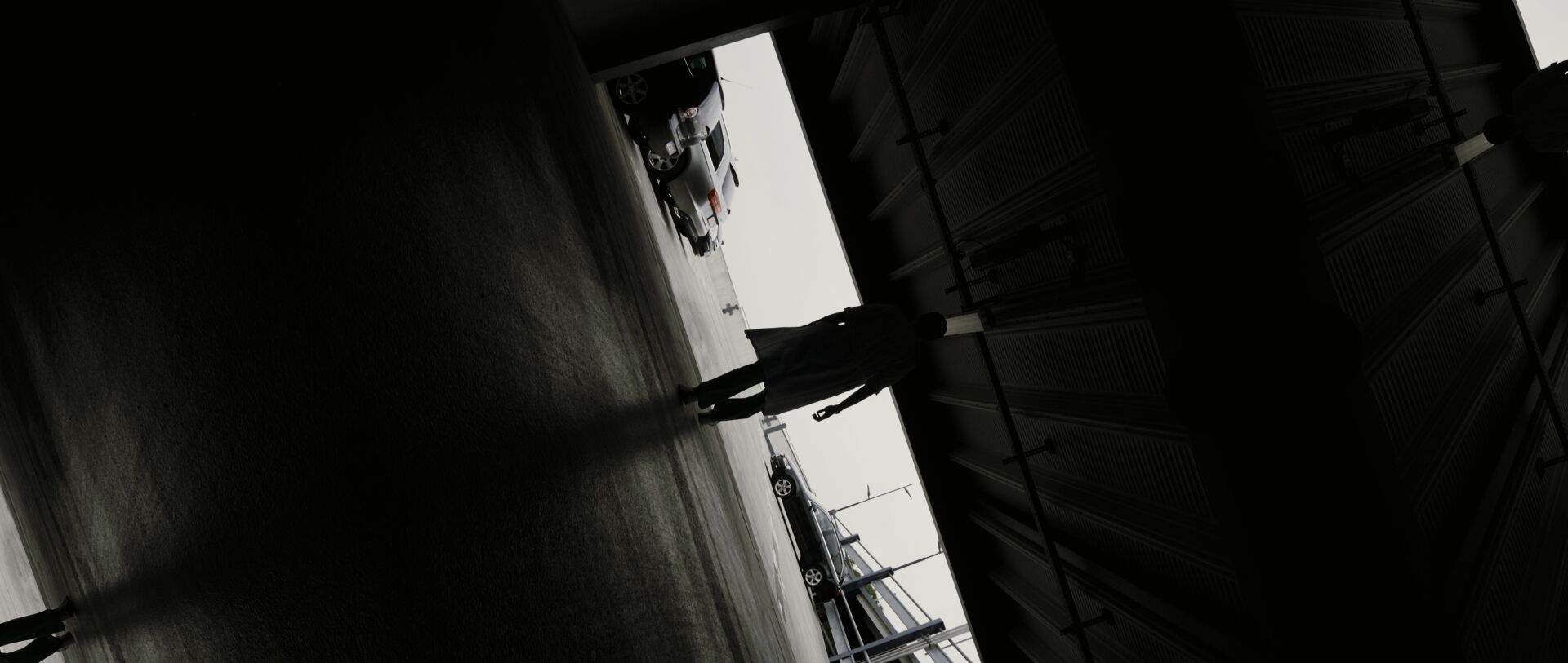 A man with a coat walks out of the dark into the light, filmed from the back. The image is rotated nearly 90 degrees to the right