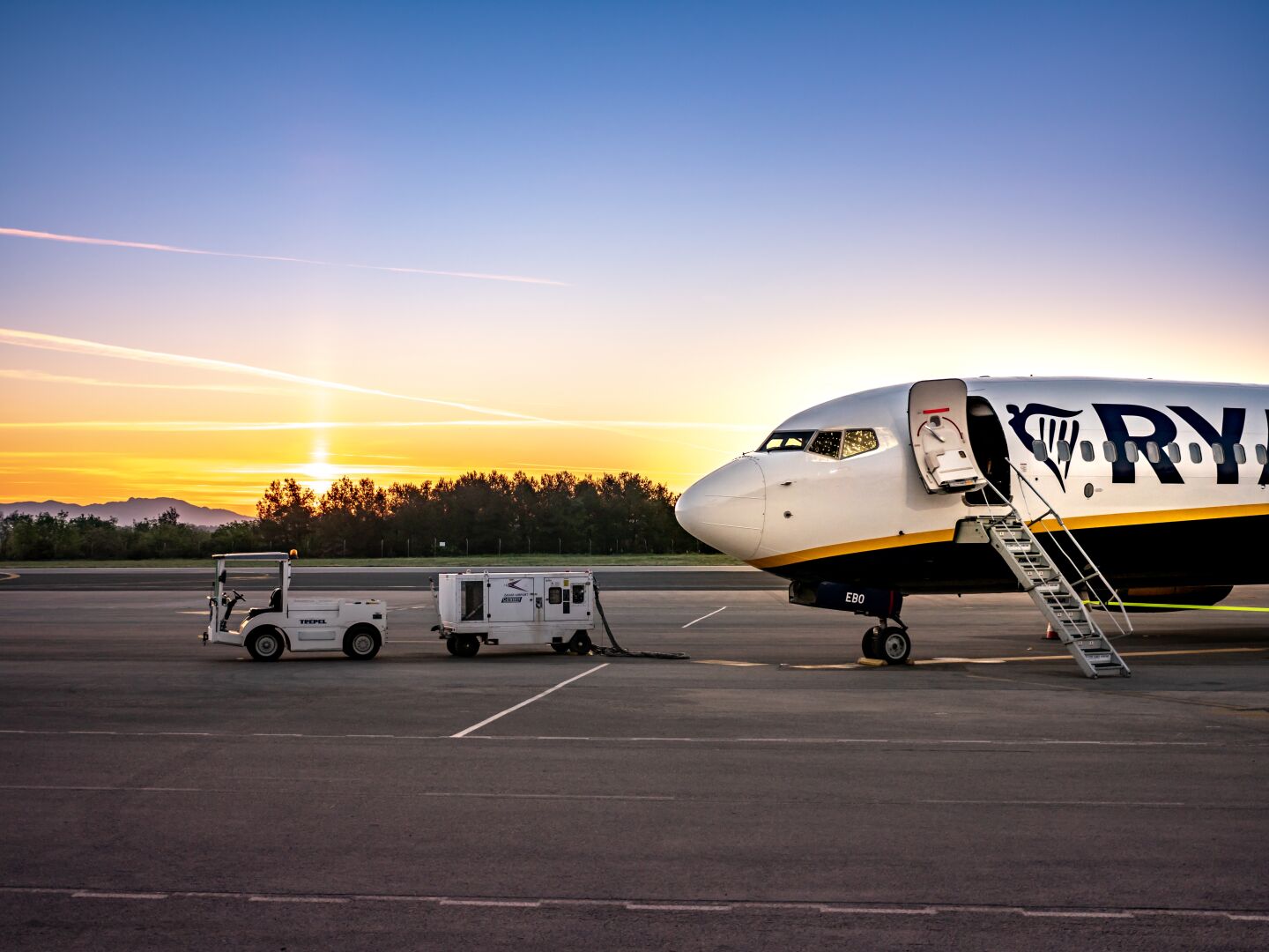 A Ryanair plane sits on the tarmac with the stairs extended and a small vehicle pulling the GPU in front. The sun is setting behind it