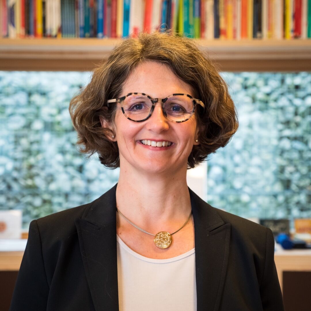 A woman in a black blazer and glasses stands in front of a window and smiles into the camera
