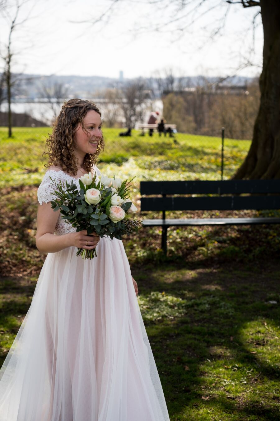 A bride stands on a meadow with the sea and a forest in the background, looking towards the right with a bouquet of flowers in her hand