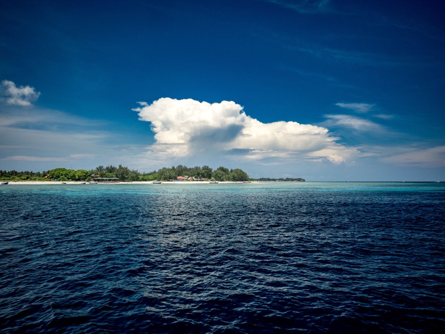 Sky and sea fight for who is bluer. In the distance a small island with white beach and palm trees, stretching into the sea, with the water showing lighter and lighter blue color the closer you get to the island