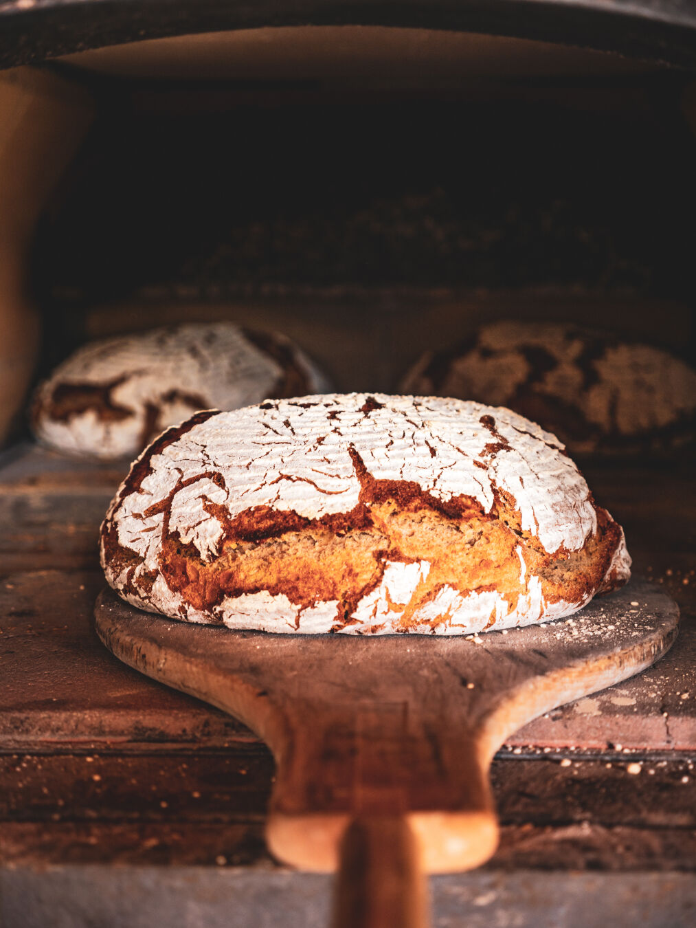 A loaf of bread while being taken from the oven still on the wooden bread puller with two more loafs in the oven behind