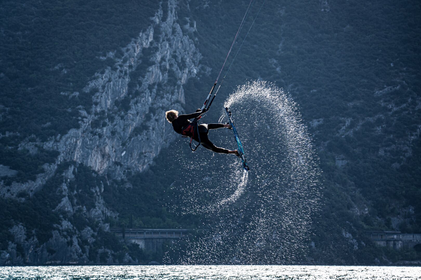 A kiter jumps up into the air while pushing his board down on one side, pulling up water and leaving a water trail in the air that looks a bit like a snail house