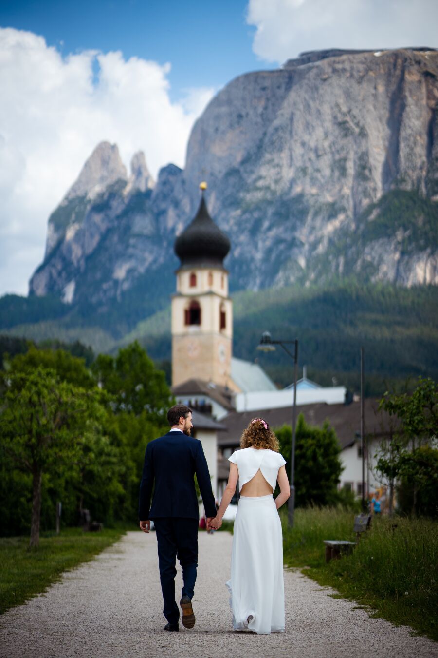 A groom and bride walk on a path holding hands towards a church in the background. Behind the church a massive mountain is visible