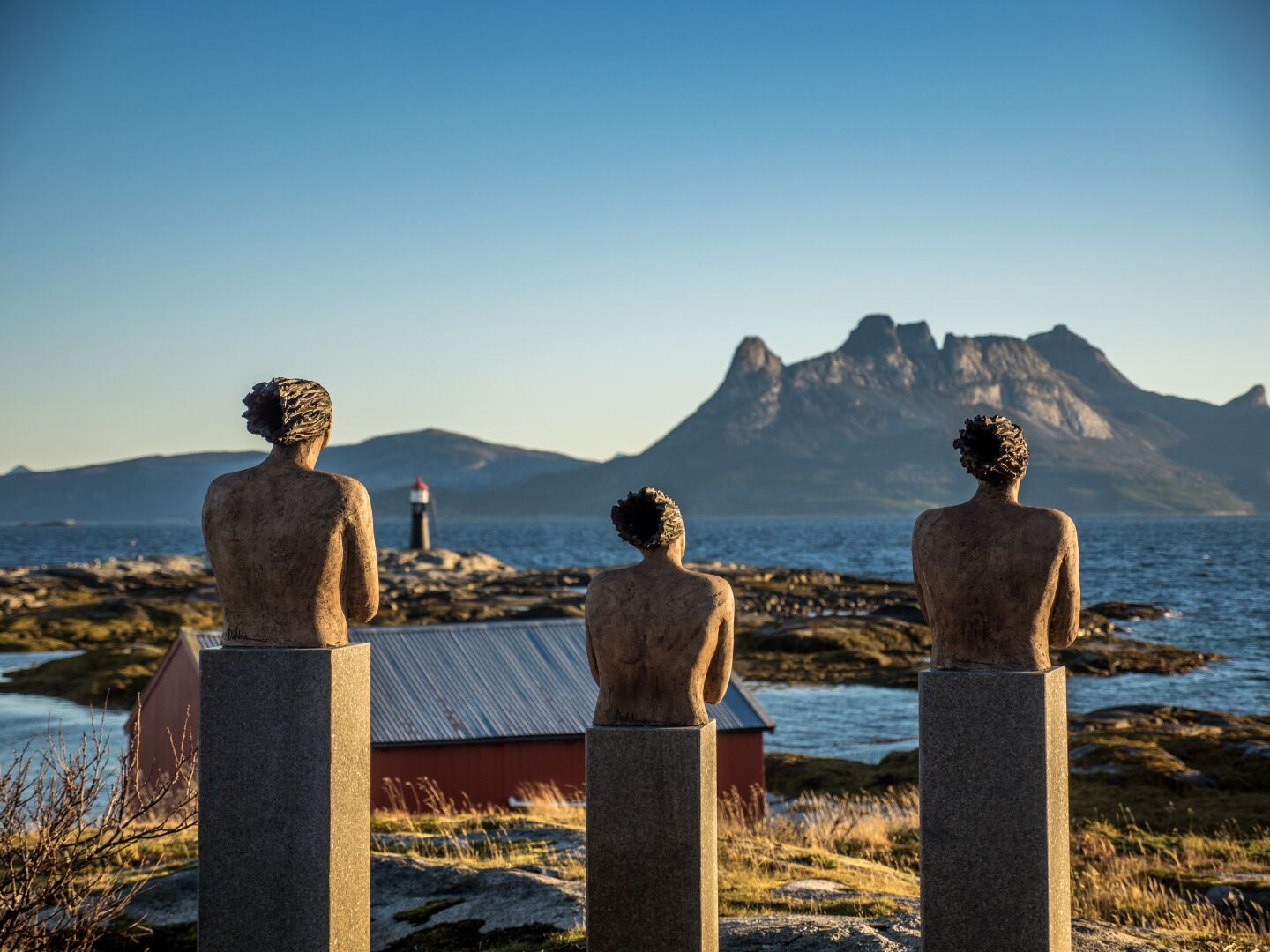 Three statues of woman torsos out of stone look away from the camera, with a red hut, the sea and the mountains on the Lofoten islands in the background