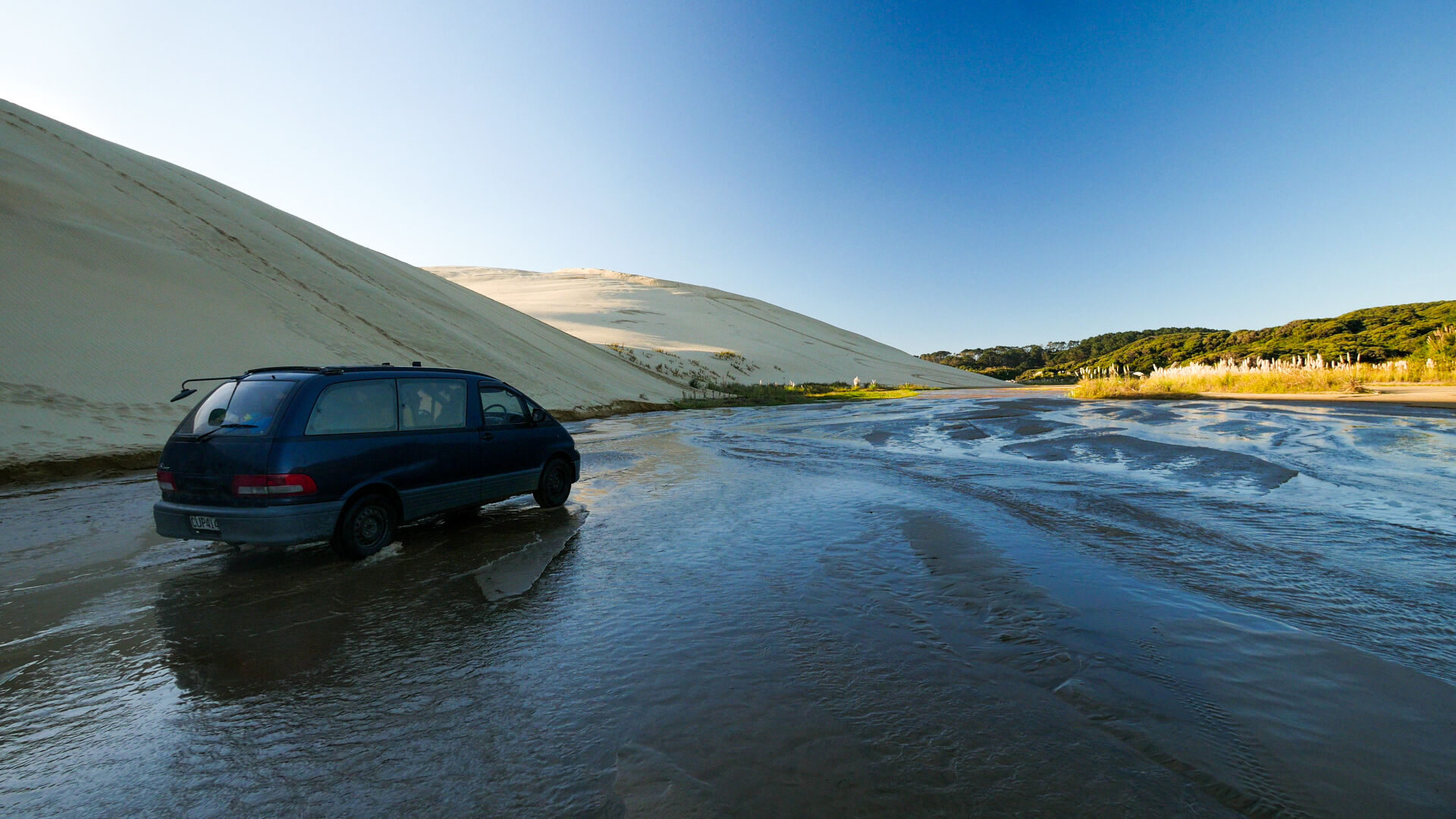 A car driving through a shallow river, with sand dunes on the left and green hills on the right