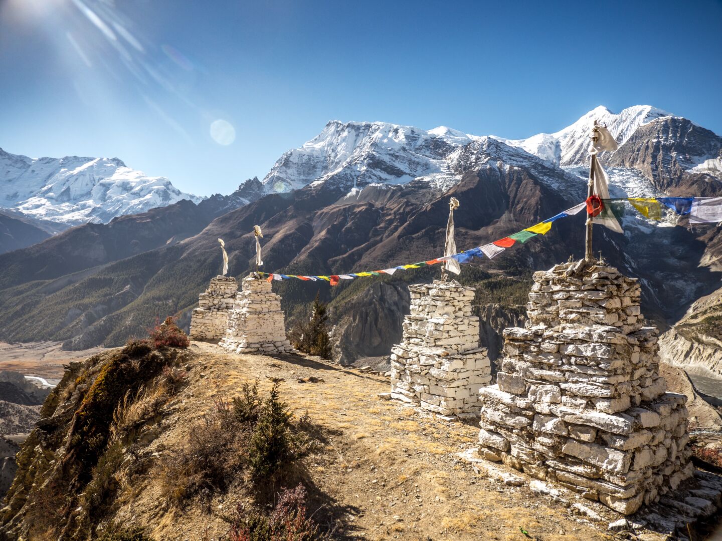 Stone pillars painted in white with colourful flags connecting all four of them. In the background white mountains growing into the sky