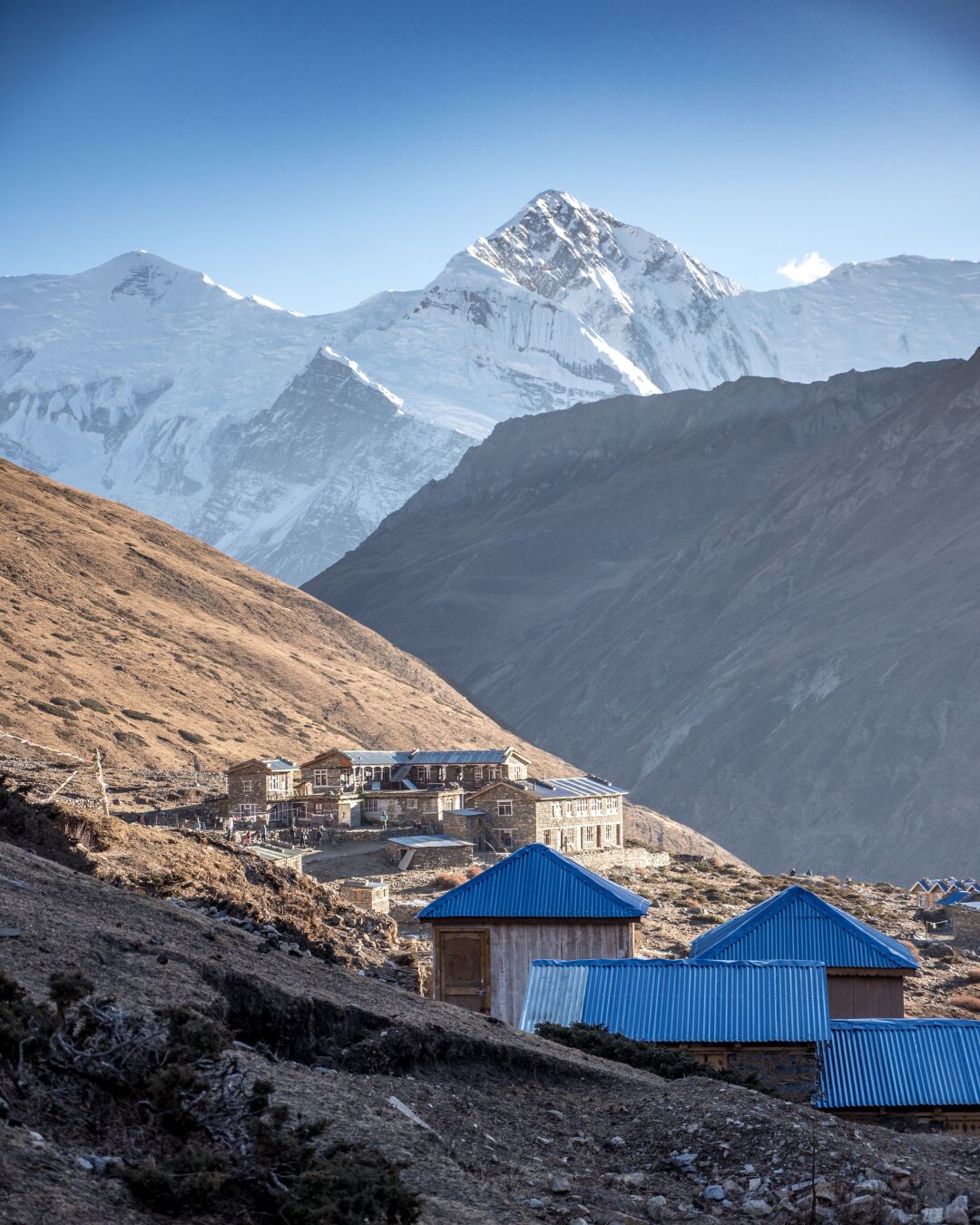 Buildings in brown camouflage on the flank of one of the mountains, with a deep valley and a snow covered mountain behind it