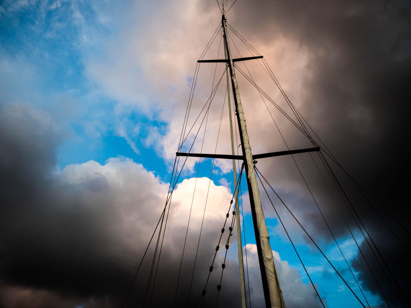 A ships mast photograph from the bottom up in front of two rather dark clouds, with a bit of blue sky left in the middle