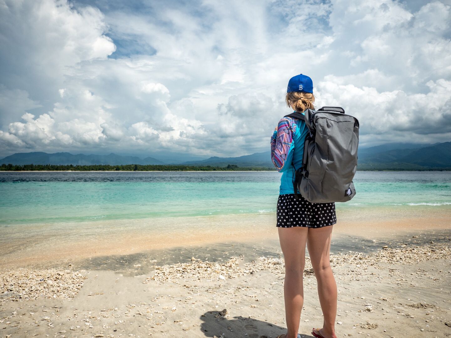 A woman in shorts with a backpack stands at the beach looking out into the very clear and blue sea