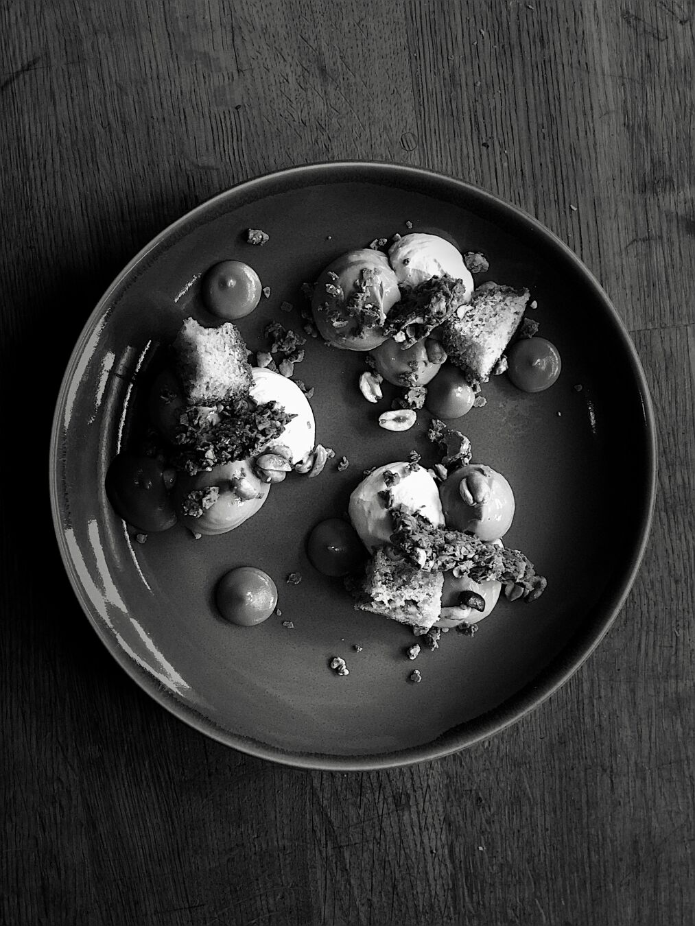 A black and white image of a plate on a wooden table with ice cream, nuts and biscuit parts arranged