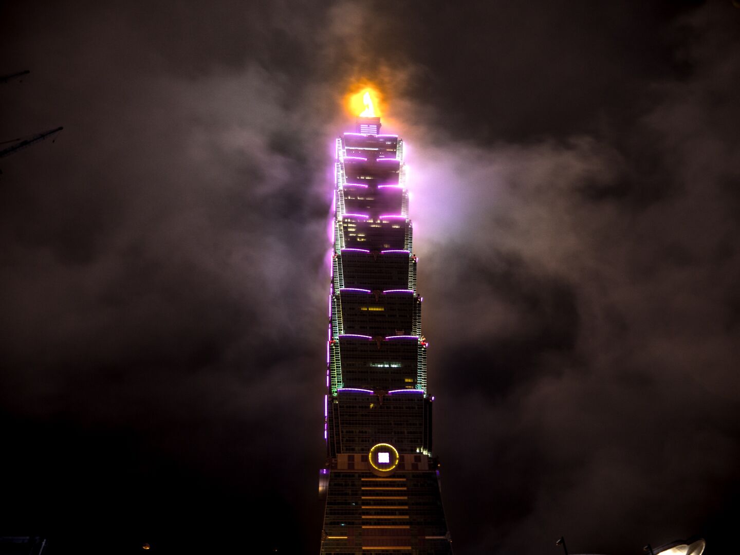A skyscraper with its top in the clouds, the corners of the building light up in pink, and the top antenna glowing in orange, lighting up the clouds around it
