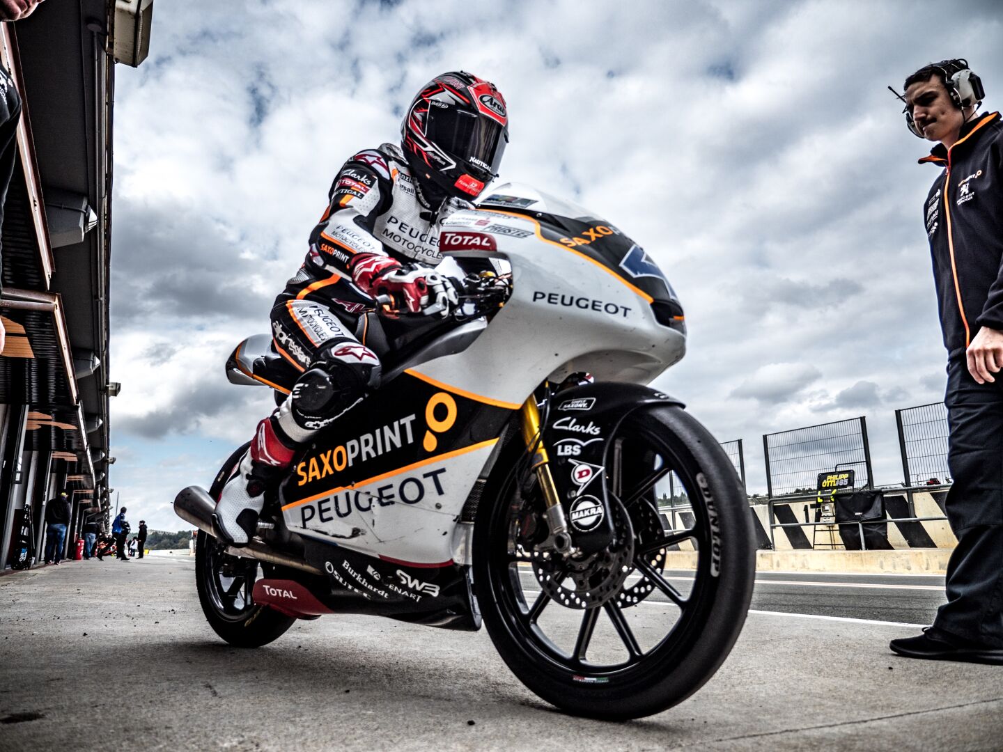 A racing driver on his racing bike ready to drive out on the track, with a mechanic with headset standing on the right side