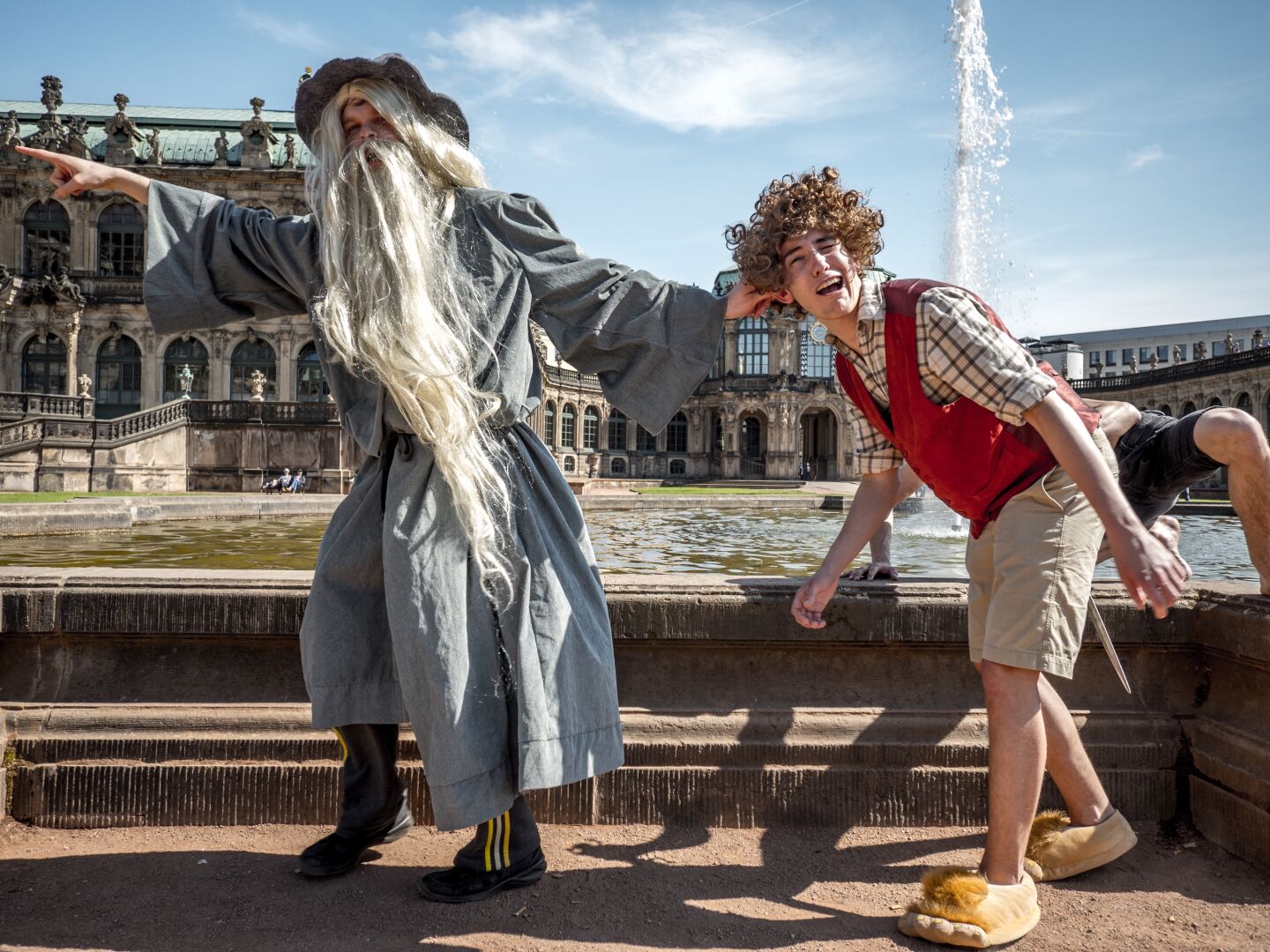 A man in a grey wizard costume pulls another man with a curly wig, a red jacket and short trousers by his ear into the left direction. Behind them a fountain and an old castle